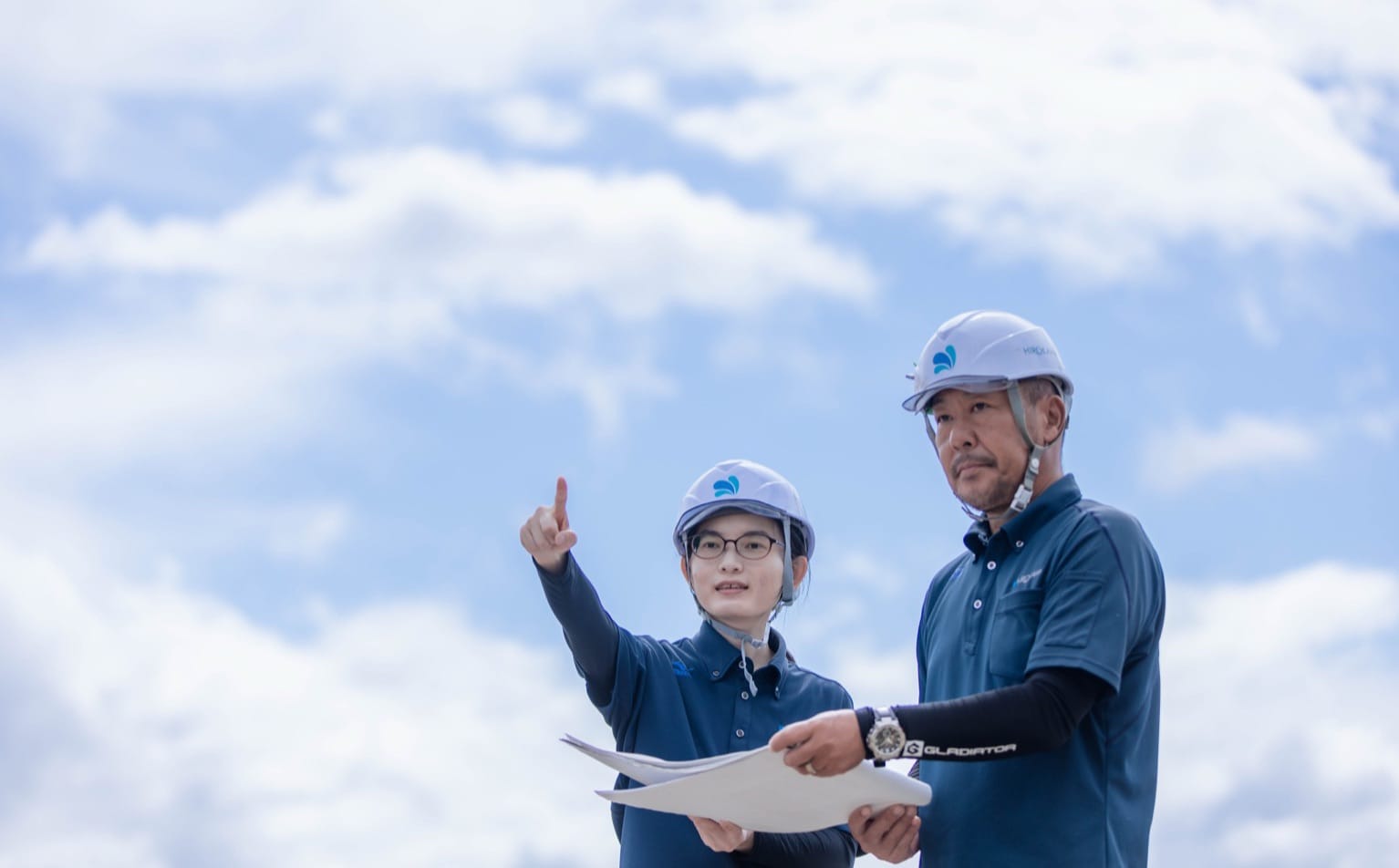 青空の下、図面を手に現場を指し示すヘルメット姿の社員