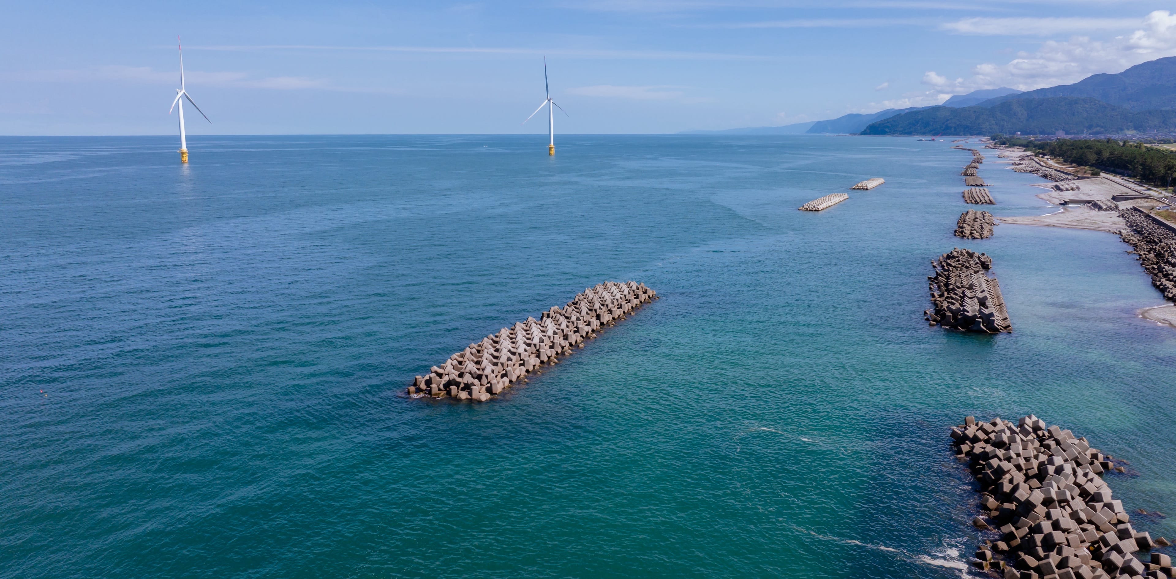 洋上風力発電の風車と消波ブロックが並ぶ海岸を上空から見た風景